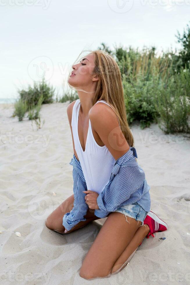 vicino su ritratto di biondo seducente ragazza con lungo brillante ondulato capelli su estate spiaggia godendo vacanza. a strisce camicetta, jeans corti. cannuccia cappello. foto