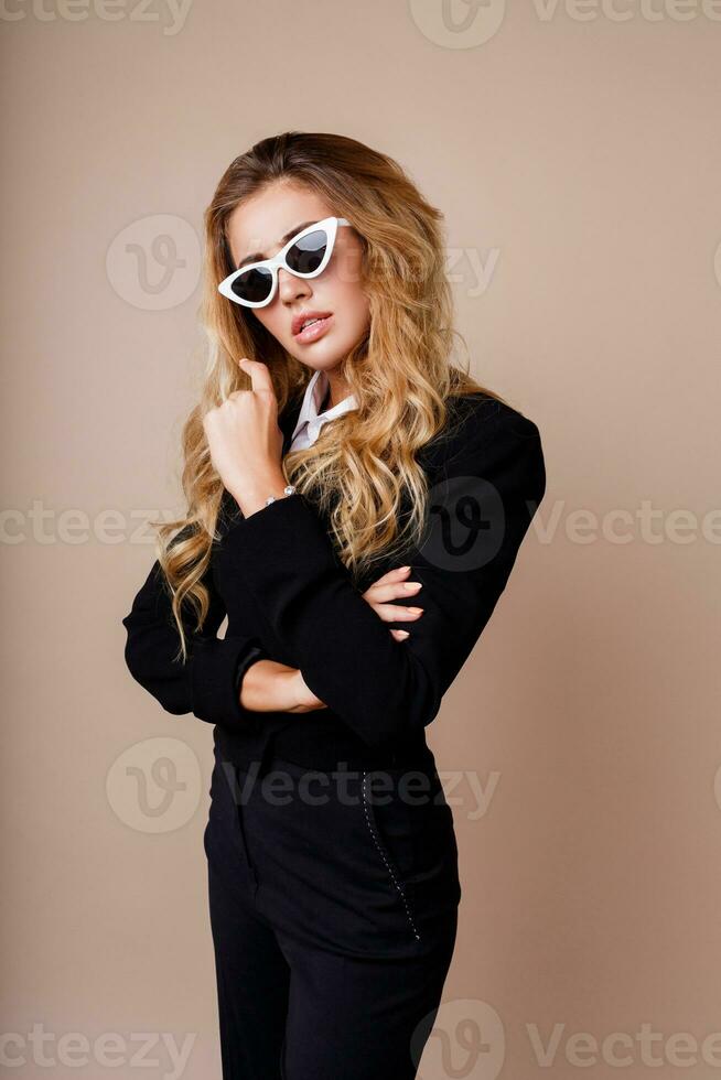 vicino su moda ritratto di bellissima biondo donna nel elegante casuale nero giacca in posa su beige sfondo. bianca retrò occhiali. alto moda Guarda. foto