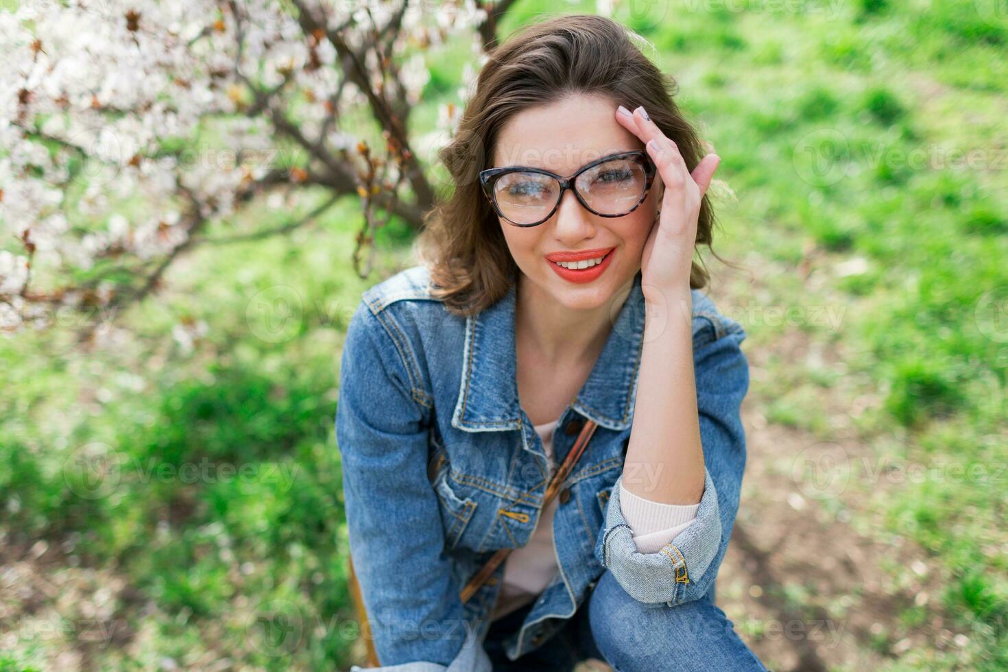 sexy donna nel jeans giacca, rosso pieno labbra, ondulato acconciatura in posa vicino fiore albero nel primavera giorno. foto