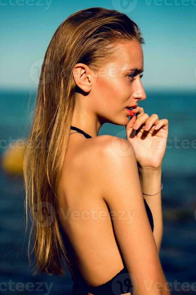 attraente giovane donna con Perfetto abbronzatura in forma corpo in posa su il spiaggia contro blu cielo. indossare costumi da bagno , bagnato capelli, naturale rendere su. foto