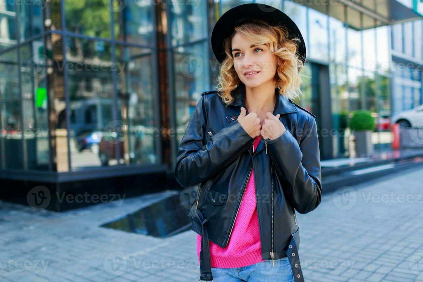 contento biondo donna in posa su moderno strade, potabile caffè o cappuccino. elegante autunno vestito, pelle giacca e a maglia maglione. urbano sfondo. rosa occhiali da sole. foto