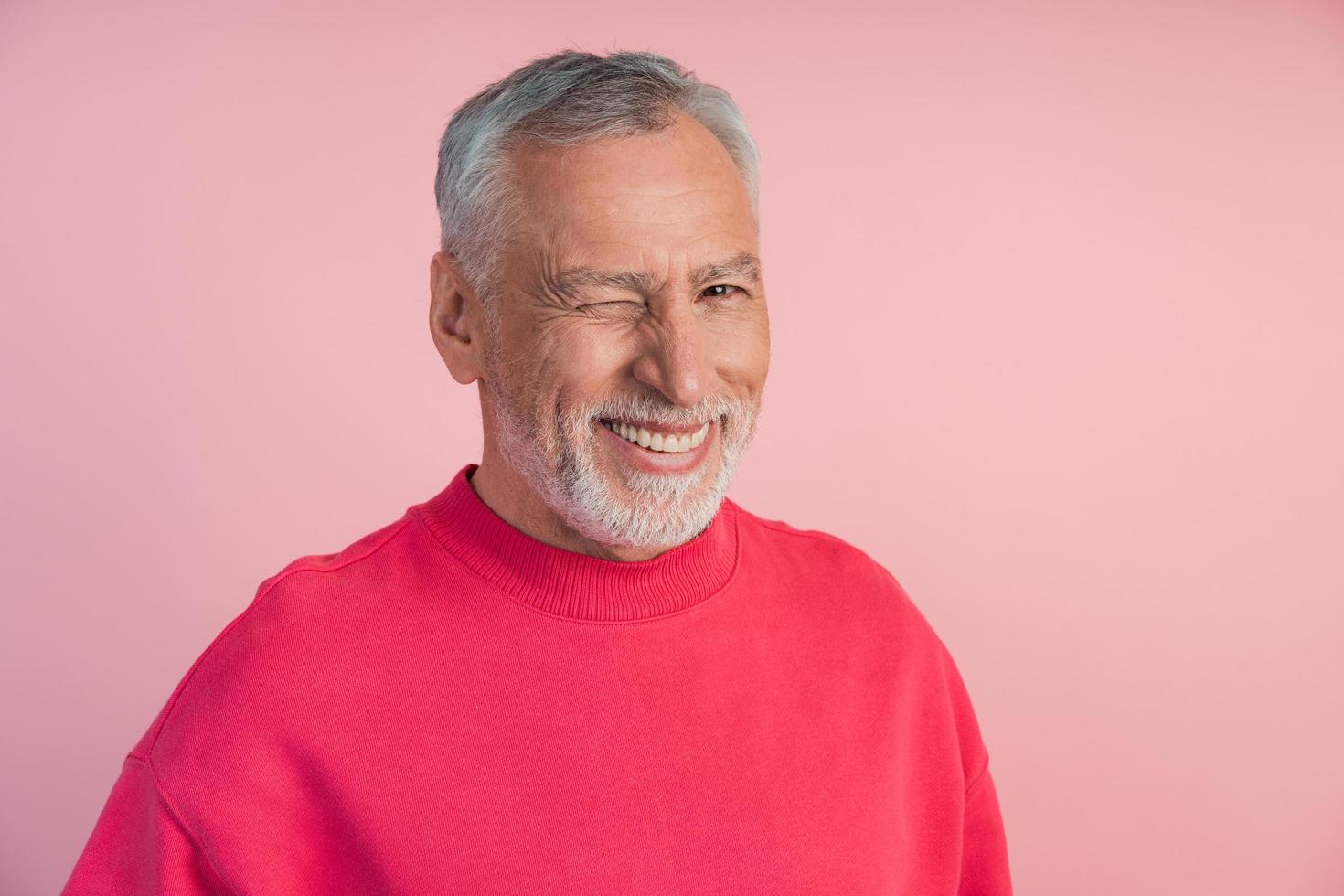 curioso, uomo anziano in un pullover luminoso strizza l'occhio su uno sfondo rosa foto
