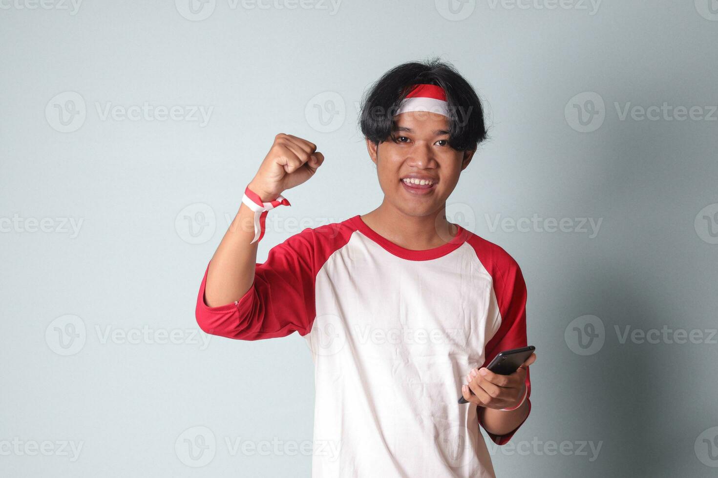 ritratto di attraente asiatico uomo nel maglietta con rosso e bianca nastro su testa, Tenere mobile Telefono mentre raccolta il suo cazzotto, festeggiare successo. isolato Immagine su grigio sfondo foto