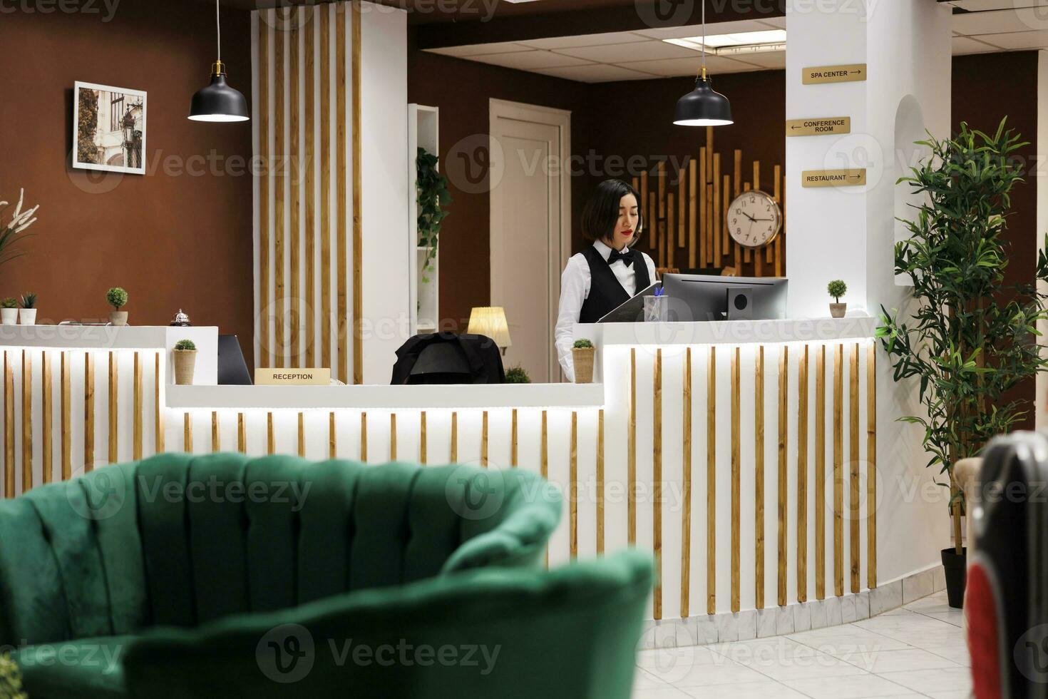 Hotel concierge scrittura prenotazione modulo a davanti scrivania, preparazione per Aiuto ospiti con alloggio a arrivo. femmina ricorrere dipendente controllo registrazioni nel disco, ricezione la zona. foto