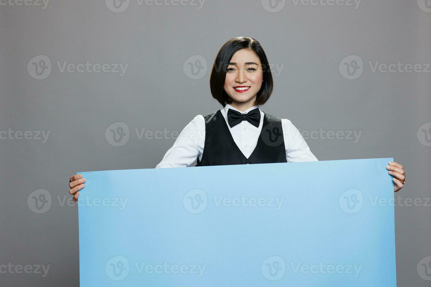 sorridente asiatico cameriera Tenere vuoto bandiera con copia spazio per ristorante pubblicità e guardare a telecamera. allegro donna addetto alla reception nel uniforme in piedi con vuoto lavagna nel mani ritratto foto