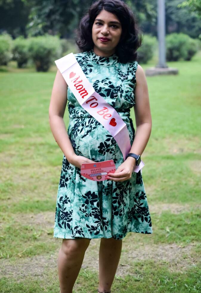 un' incinta indiano signora pose per all'aperto gravidanza sparare e mani su gonfiarsi, indiano incinta donna mette sua mano su sua stomaco con un' maternità vestito a società parco, incinta al di fuori maternità sparare foto