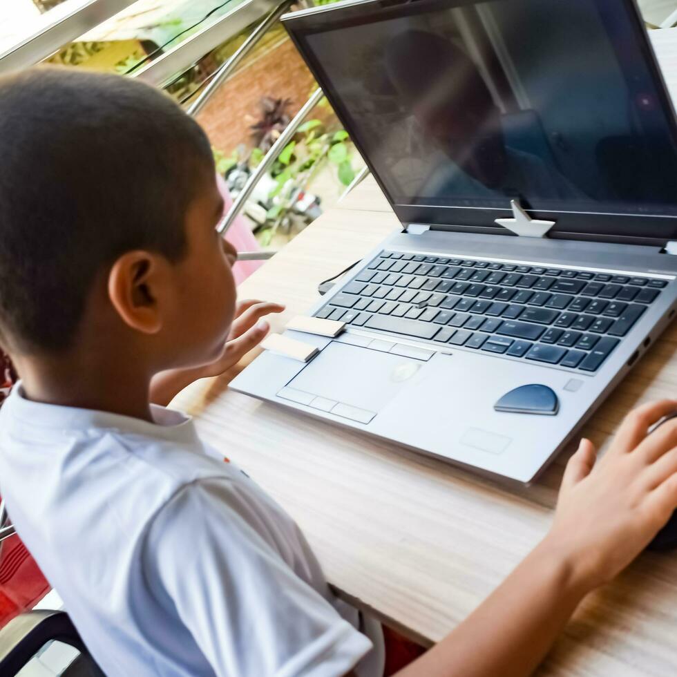 poco ragazzo seduta a tavolo utilizzando il computer portatile per in linea classe nel grado 1, bambino studiando su il computer portatile a partire dal casa per distanza apprendimento in linea formazione scolastica, scuola ragazzo bambini stile di vita concetto foto