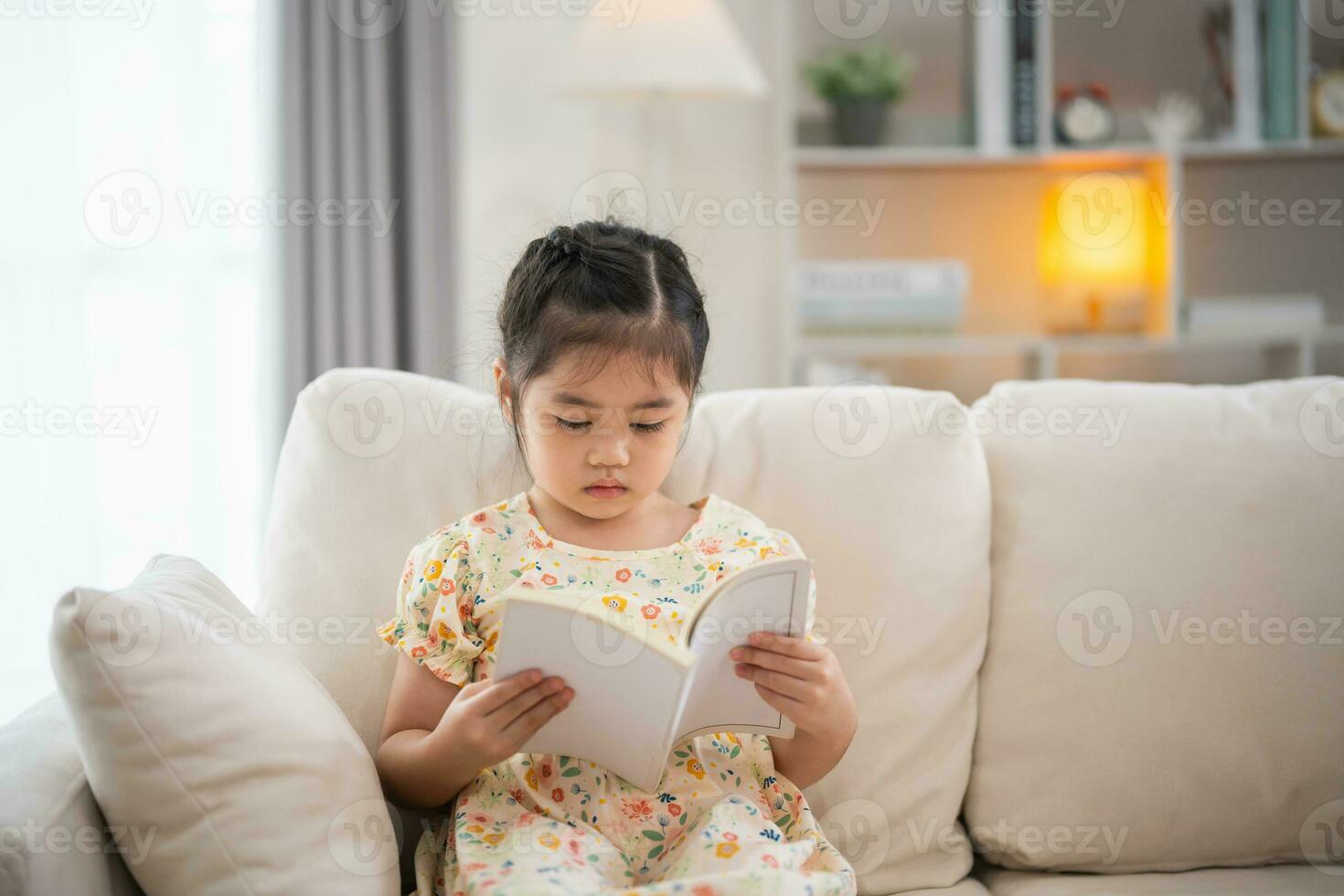 gioioso contento asiatico bambino bambino ragazza sorridente e lettura libro mentre seduta su divano divano nel vivente camera a casa. ragazza rilassati lettura libro Sorridi a divano nel il Casa. indietro per scuola concetto. foto