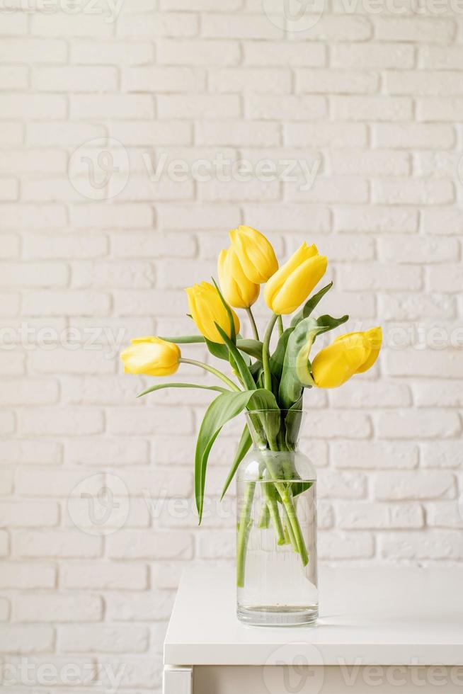 tulipani gialli in un vaso di vetro su uno sfondo di muro di mattoni bianchi foto