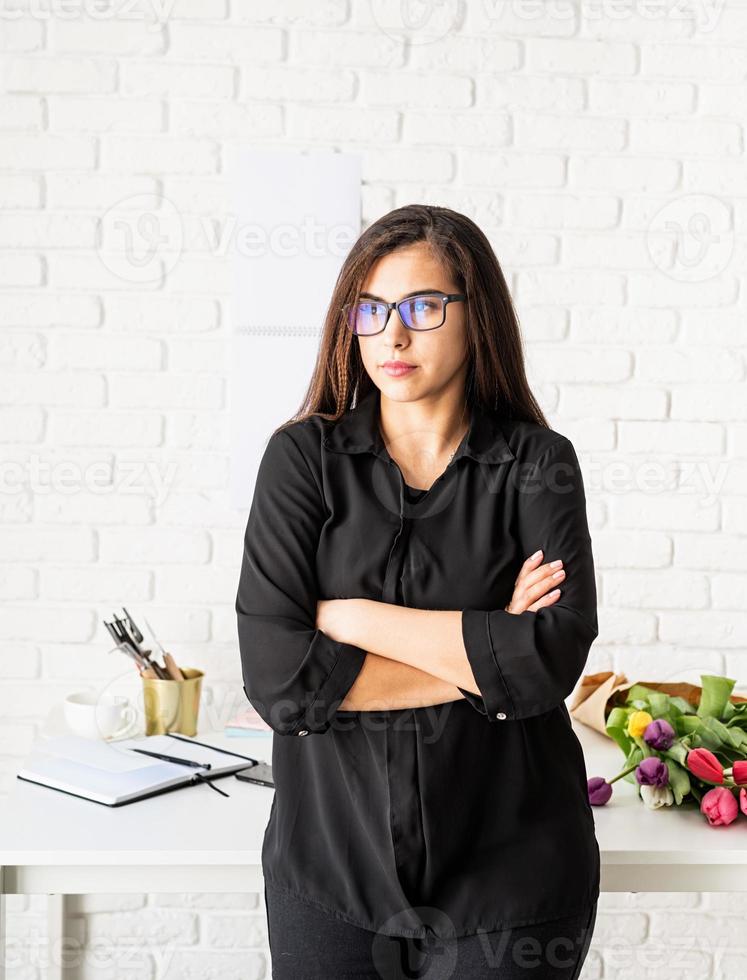 donna che lavora in ufficio, in piedi accanto alla scrivania con le braccia incrociate foto