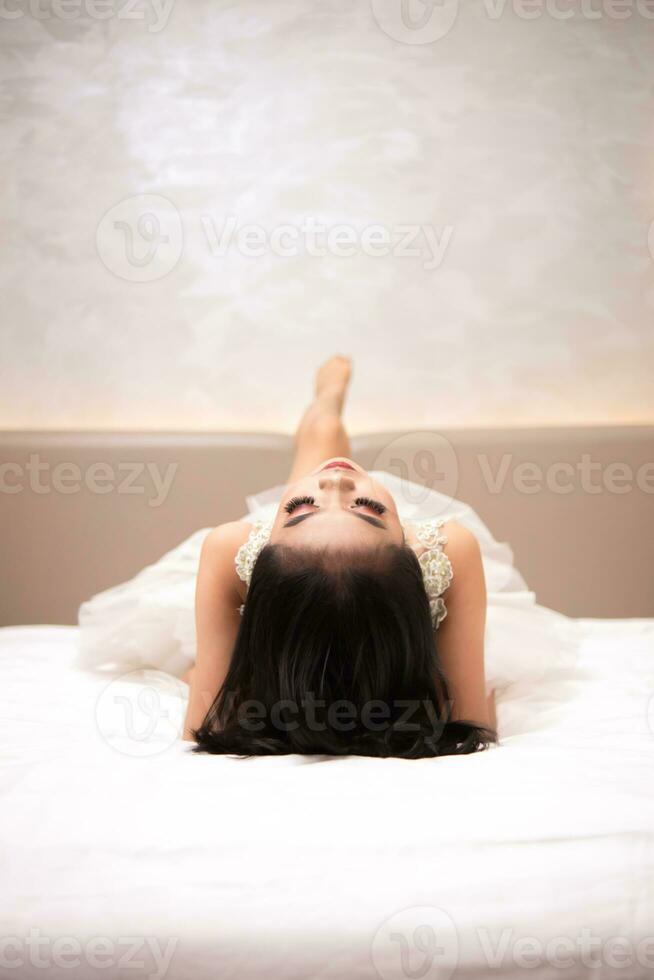un asiatico donna nel un' bianca vestito è dire bugie su un' bianca letto con sua nero capelli perdere nel un' Hotel foto