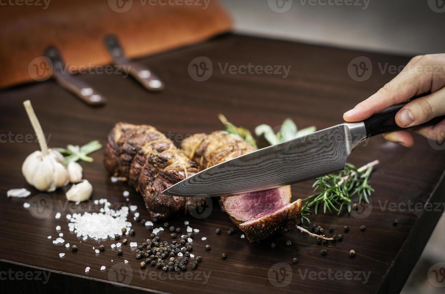 Affettare una porzione di organic roast beef roll con coltello sul tavolo di legno con aglio pepe e sale a Melbourne in Australia foto