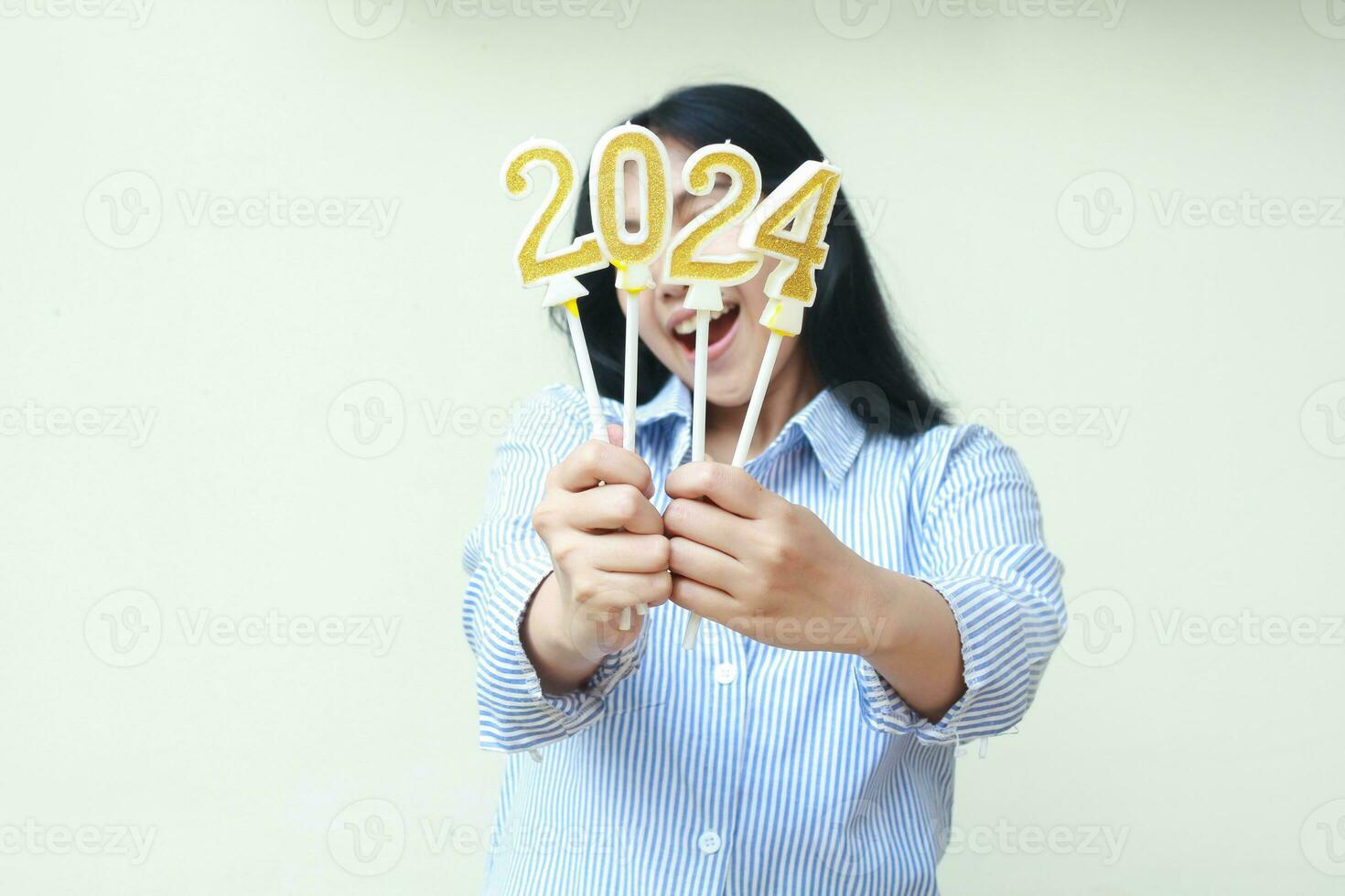 contento giovane donna asiatico mostrando d'oro candele numeri 2024 copertura viso per annunciare annuale scambio anno usurai blu strisce camicia casuale isolato foto