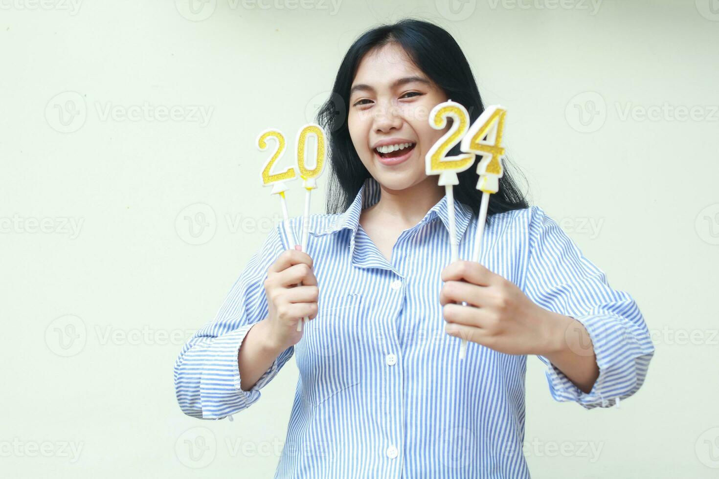 eccitato asiatico giovane donna collaboratore ridendo felicità festeggiare nuovo anni vigilia di Tenere d'oro candele numeri 2024 indossare blu strisce camicia casuale guardare a telecamera isolato su bianca sfondo foto