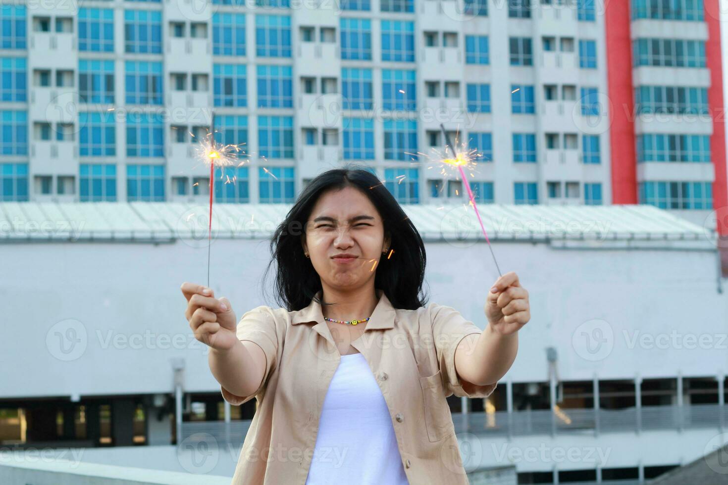 contento asiatico giovane donna fricchettone Tenere sparklers fuoco d'artificio celebrare nuovo anno vigilia con danza nel tetto superiore appartamento con urbano edificio sfondo foto