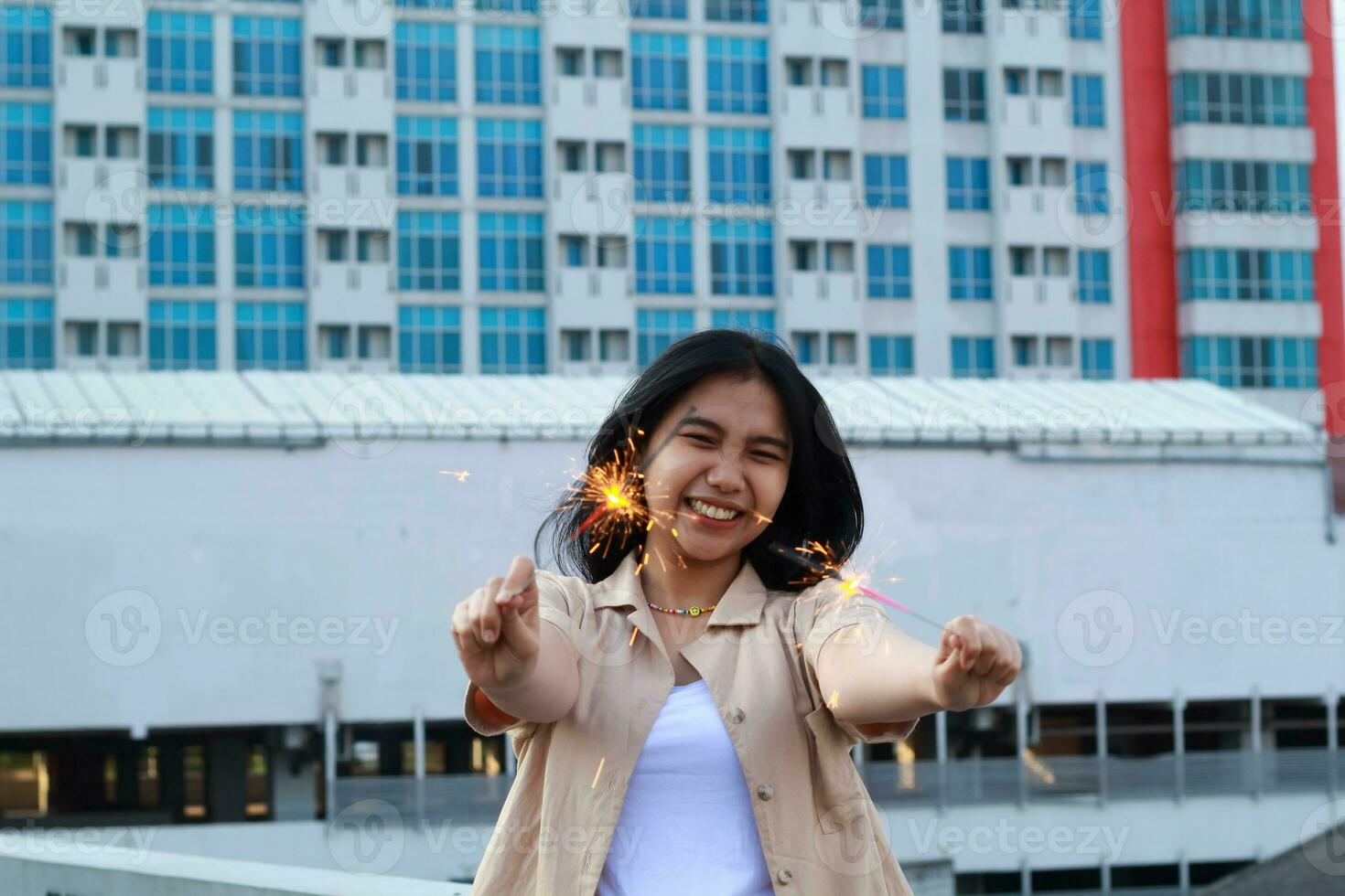 spensierato asiatico giovane donna Tenere sparkler celebrare nuovo anno vigilia con ridere e danza nel tetto appartamento all'aperto con città edificio sfondo foto