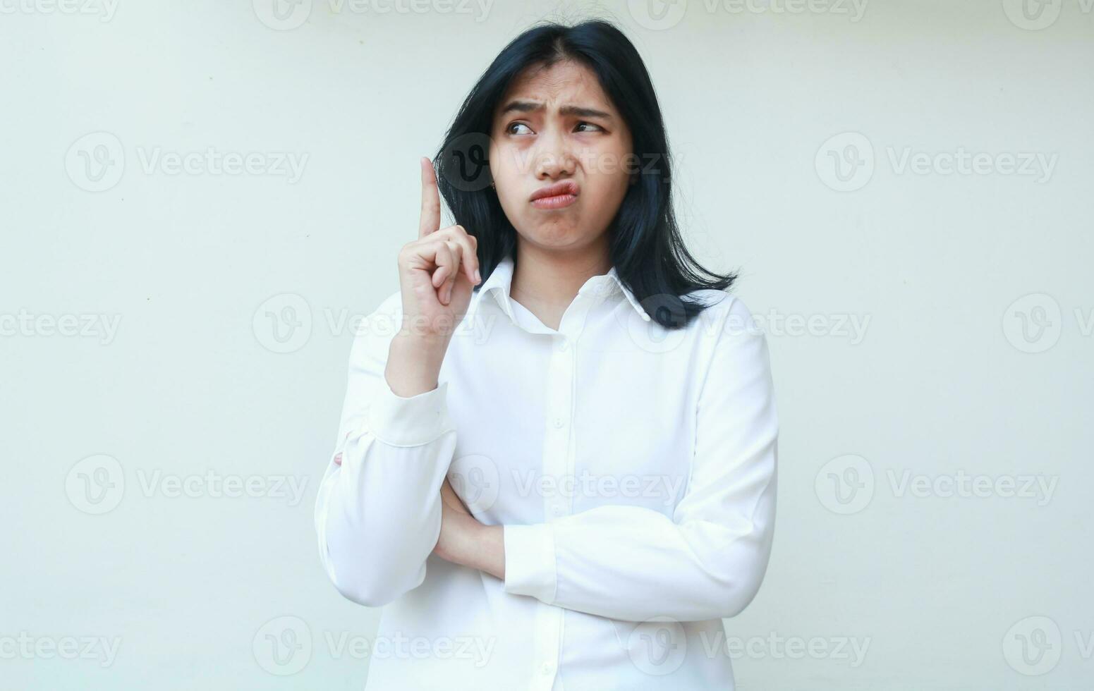 ritratto di riflessivo asiatico giovane donna ufficio dipendente guardare sopra con curiosità e puntamento su fare presentazione gesto con piegato braccio, in piedi confuso foto