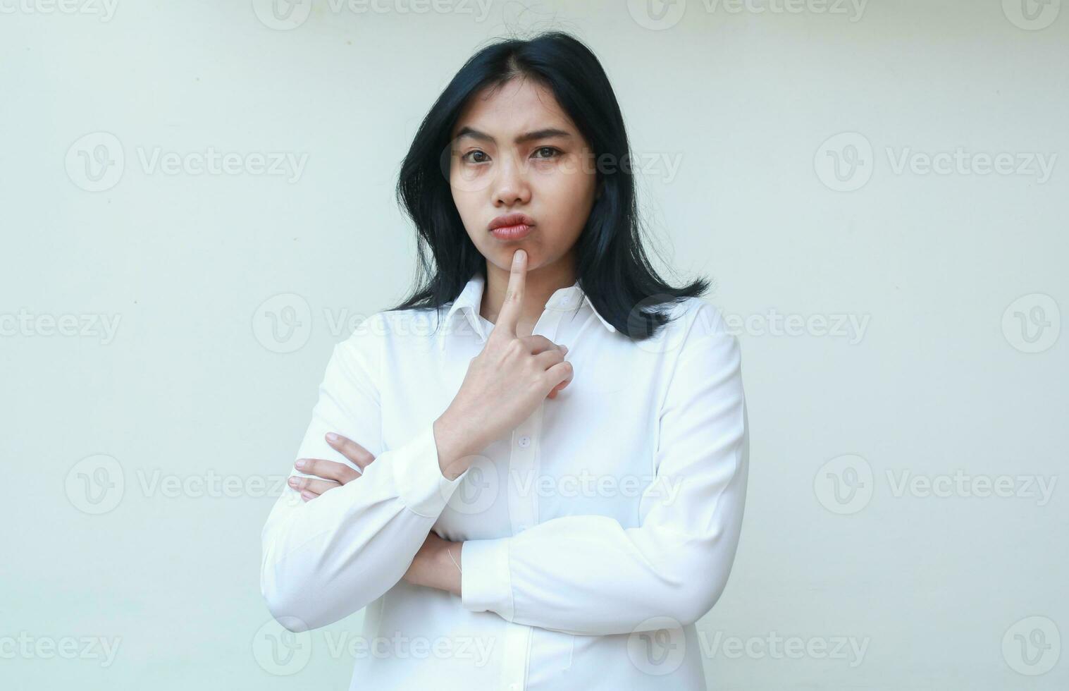 Immagine di pensieroso bella asiatico donna manager indossare bianca camicia guardare a telecamera meditando con dita su mento e piegato braccio, in piedi insicuro espressione foto