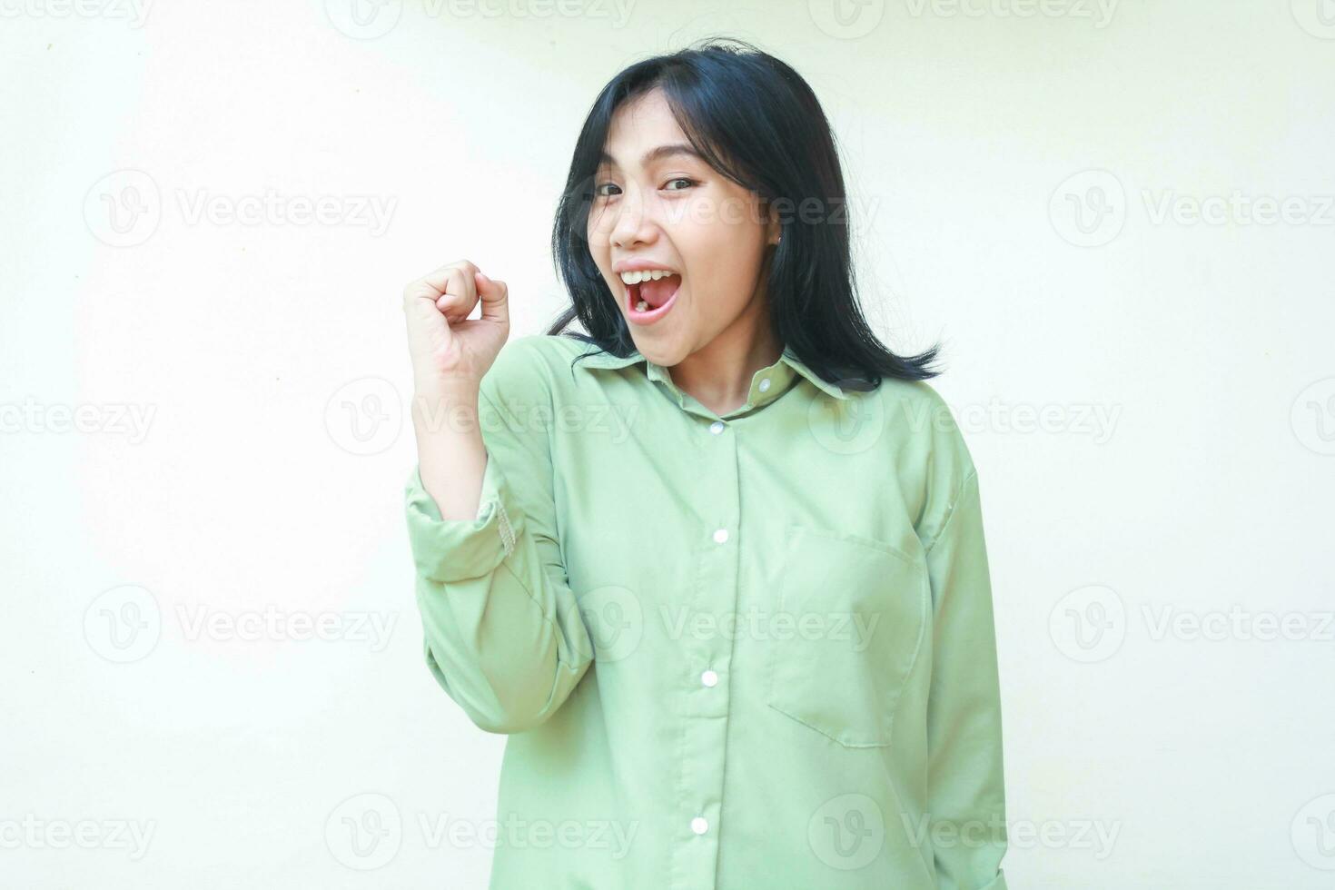 giocoso successo buio capelli asiatico donna ridendo e danza per celebrare campione vincere vittoria di aumentare cazzotto indossare verde oversize camicia in piedi al di sopra di isolato bianca sfondo, guardare a telecamera foto