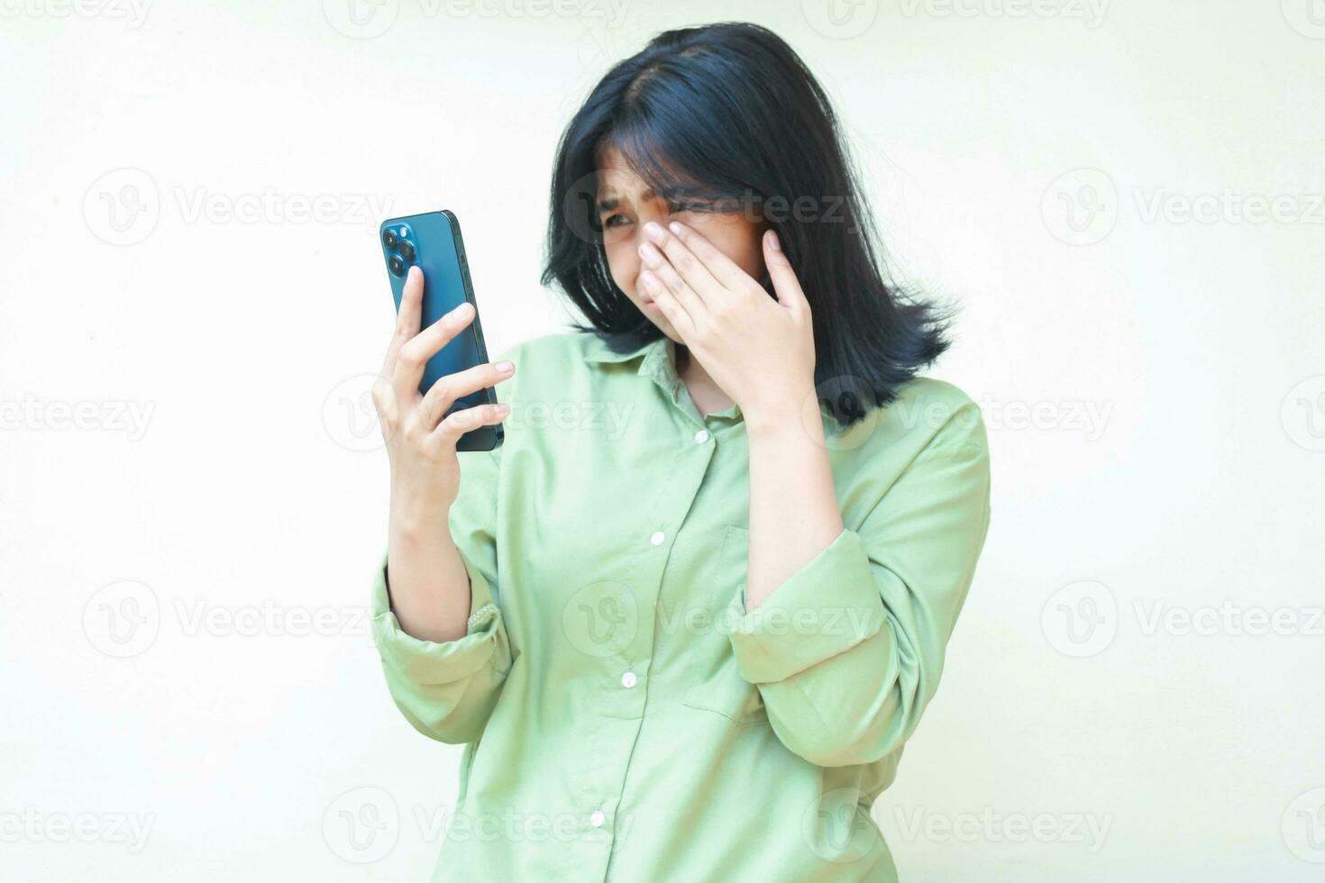 triste asiatico donna salviette sua lacrime dopo piangere per utilizzando sociale media su smartphone hold su mani indossare verde sovradimensionato camicia in piedi al di sopra di isolato sfondo. mentale Salute concetto, bullismo foto