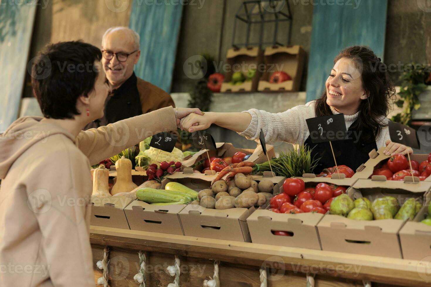 sorridente donna Locale venditore in piedi dietro a In piedi con fresco biologico produrre saluto regolare cliente, amichevole femmina contadino tremante mano di consumatore mentre vendita homegrown cibo a azienda agricola mercato foto