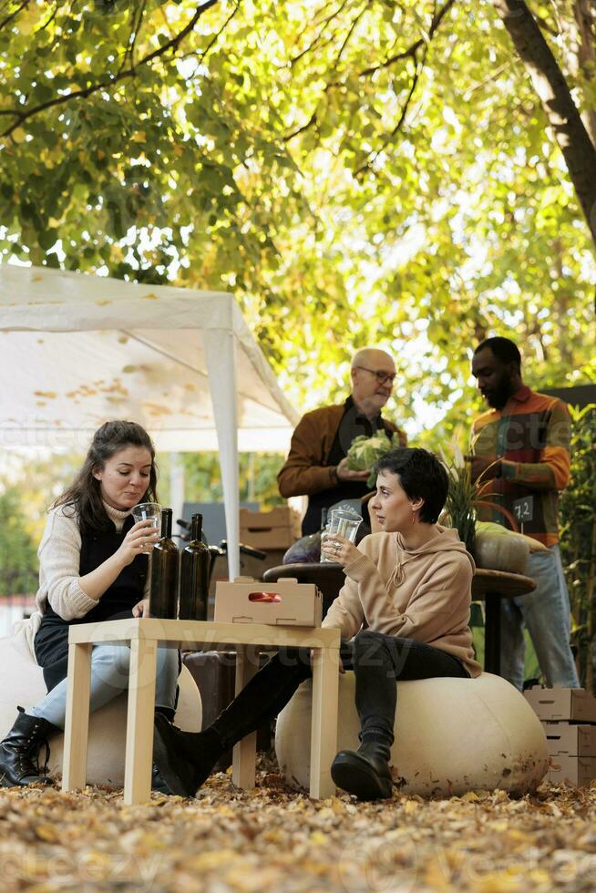 giovane donna cliente seduta a tavolo con Locale enologo degustazione fatti in casa naturale vino prima acquisto mentre shopping a biologico cibo Festival. Prodotto campionamento a agricoltori mercato foto