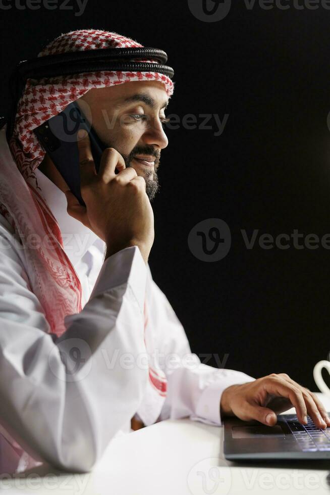 mezzo orientale uomo, vestito tradizionalmente, impegnato nel un' Telefono conversazione, lui multitasking con il suo mobile dispositivo e il computer portatile. un' moderno miscela di eredità e tecnologia. vista laterale, avvicinamento. foto