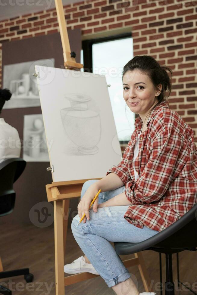 contento giovane donna sorridente a telecamera mentre seduta a cavalletto durante divertimento e creativo arte classe, apprendimento le basi di disegno, provando qualcosa nuovo. alunno ragazza godendo arti attività foto