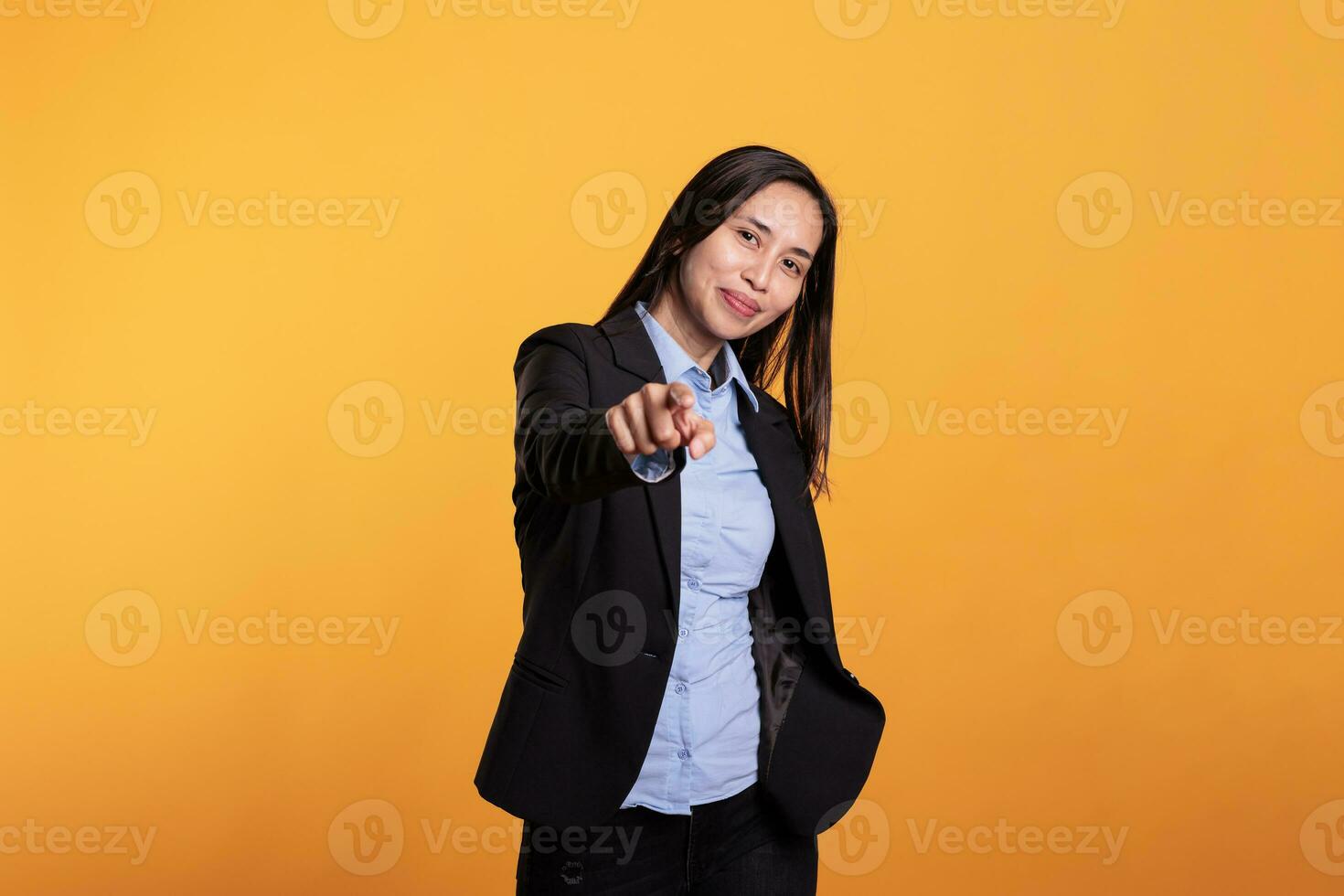 sorridente asiatico donna nel formale completo da uomo puntamento a telecamera con dita durante sparare volta. attraente giovane adulto con casuale atteggiamento utilizzando carisma, in piedi nel studio al di sopra di giallo sfondo foto
