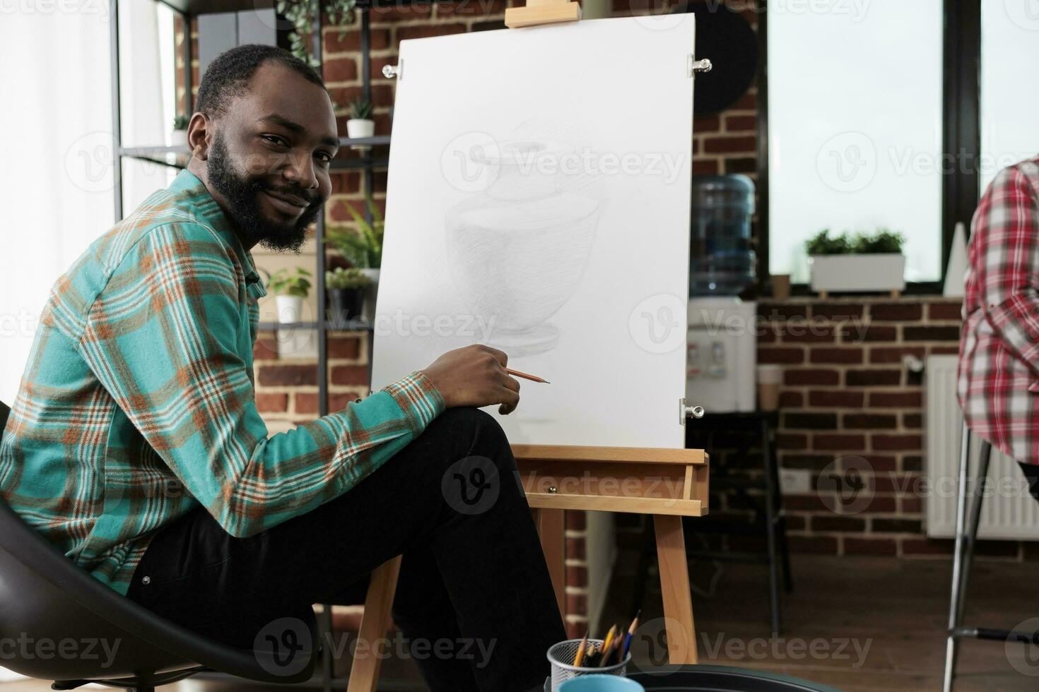 arte attività e benessere. ritratto di contento sorridente africano americano tipo durante disegno lezione, giovane nero uomo seduta a cavalletto guardare a telecamera, sensazione ispirato durante schizzi classe foto