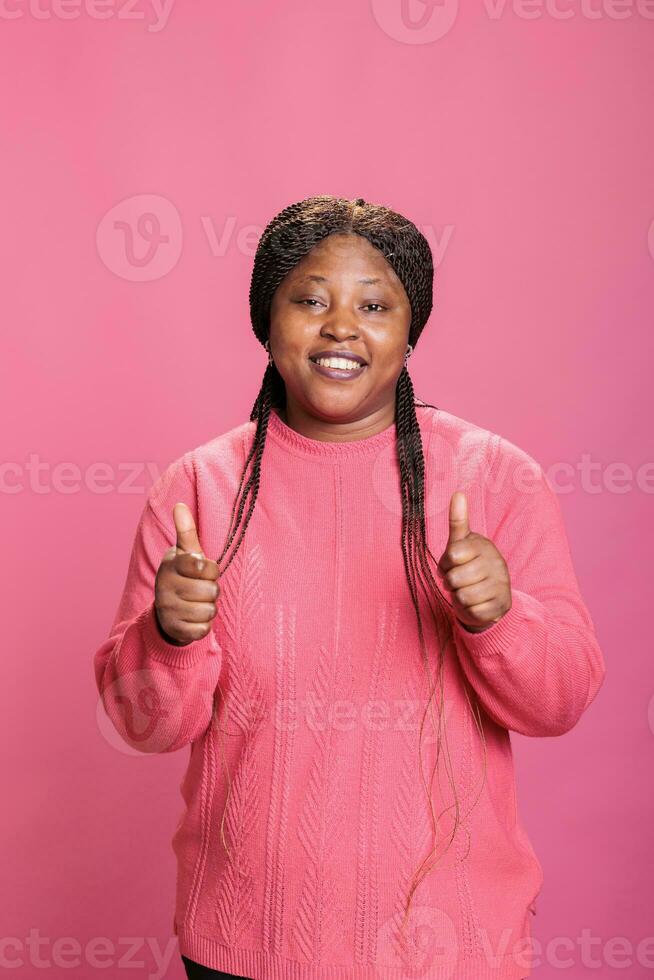 sorridente africano americano modello mostrando pollici su cartello in piedi al di sopra di rosa sfondo nel studio. contento allegro donna con gioioso facciale espressione dando approvazione gesto, persona fare piace cartello foto