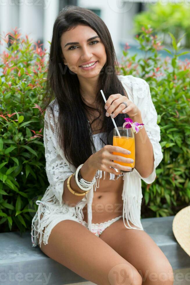 contento giovane bellissimo concia donna nel elegante boho bianca beachwear seduta vicino tropicale piscina nel lusso Hotel e godendo arancia cocktail o succo. spiaggia festa. foto