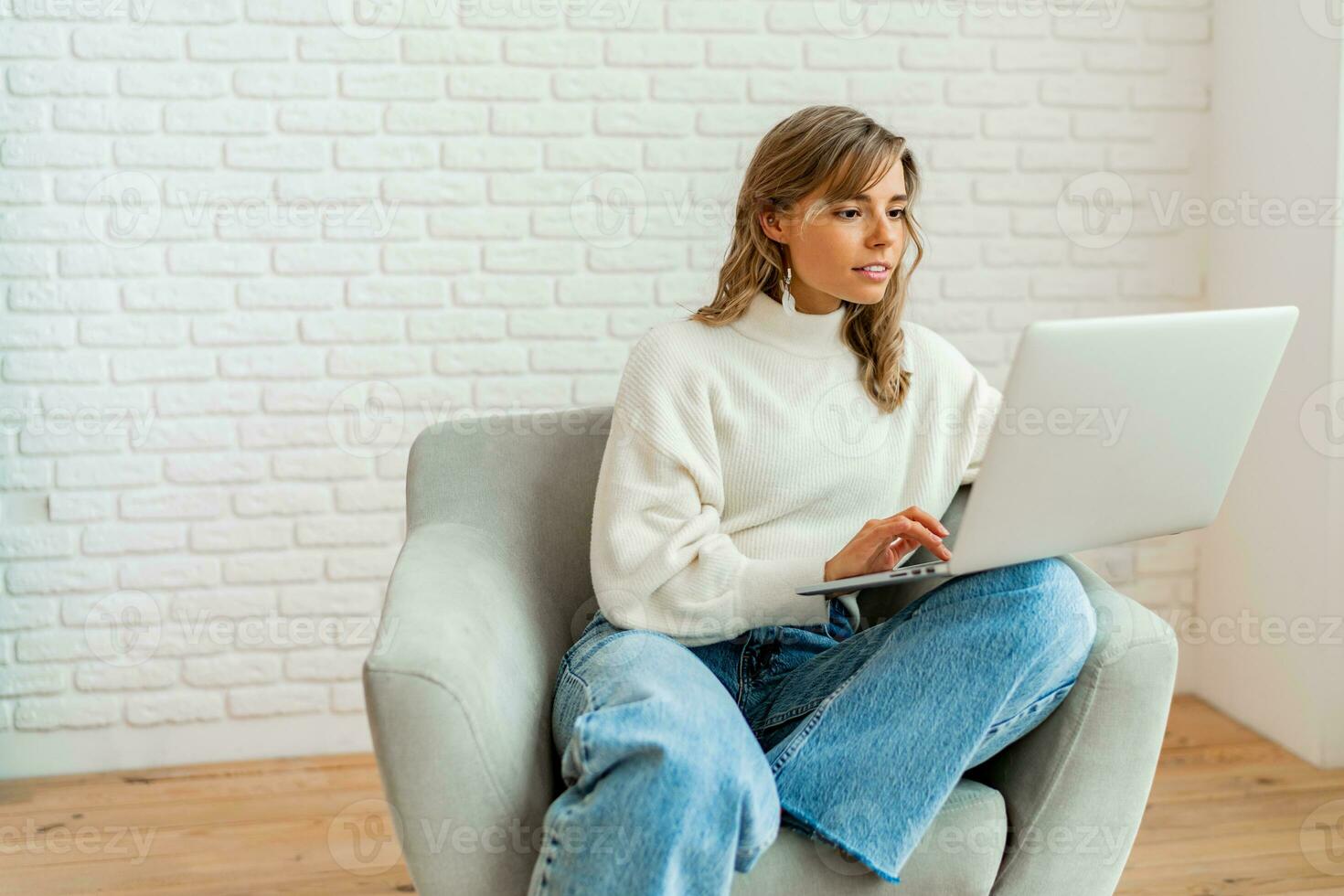 bella donna con biondo ondulato capelli seduta su divano a casa Lavorando su il computer portatile computer. indossare accogliente maglione. foto