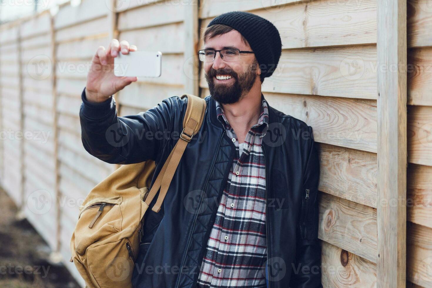 fricchettone uomo fabbricazione autoscatto su smartphoneby mobile Telefono su legna parete sfondo. indossare elegante nero cappello, pelle giacca e zaino. foto