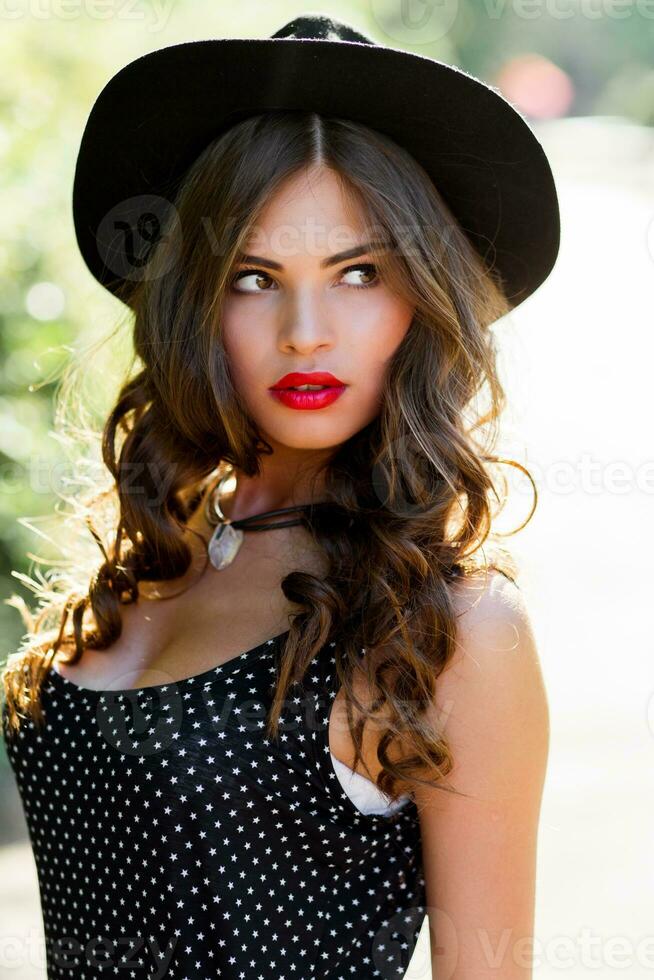 estate moda ritratto di r elegante donna con Perfetto ondulato capelli nel elegante elegante nero cappello e luminosa rendere su in posa nel il parco. strada stile. foto