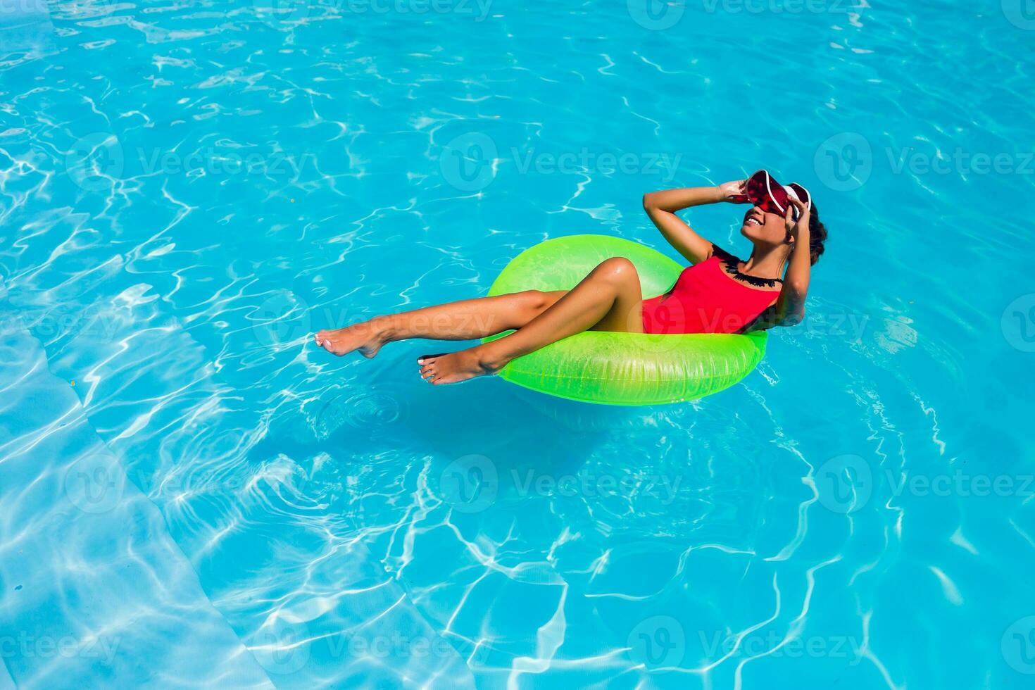 sexy abbronzato donna nel rosso costume da bagno avendo divertimento e godendo estate nel sorprendente grande nuoto piscina. giovane bella ragazza nuoto su gonfiabile squillo. elegante trasparente cap. spiaggia festa. foto