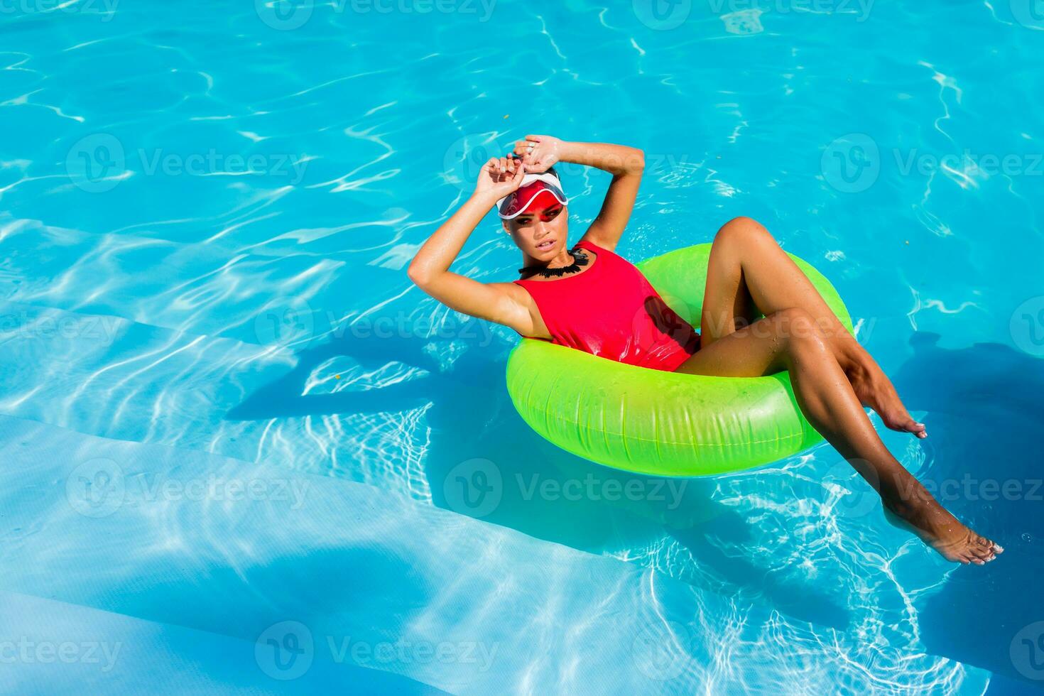 sexy abbronzato donna nel rosso costume da bagno avendo divertimento e godendo estate nel sorprendente grande nuoto piscina. giovane bella ragazza nuoto su gonfiabile squillo. elegante trasparente cap. spiaggia festa. foto