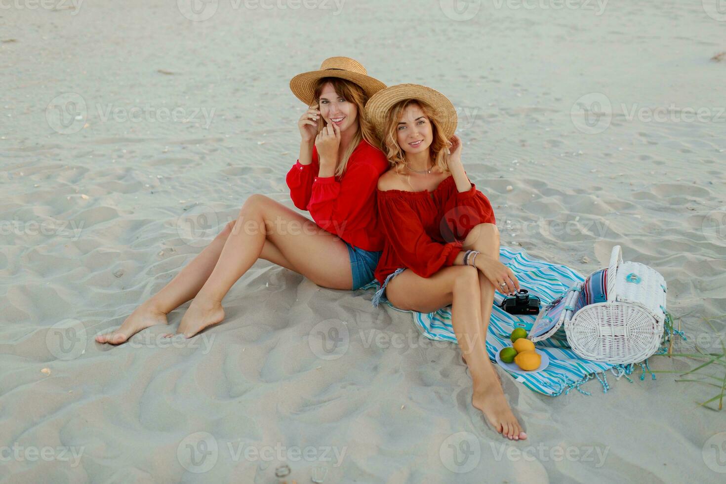 Due bella donne nel rosso estate attrezzatura abd cannuccia cappelli godendo picnic su il spiaggia. estate umore. foto