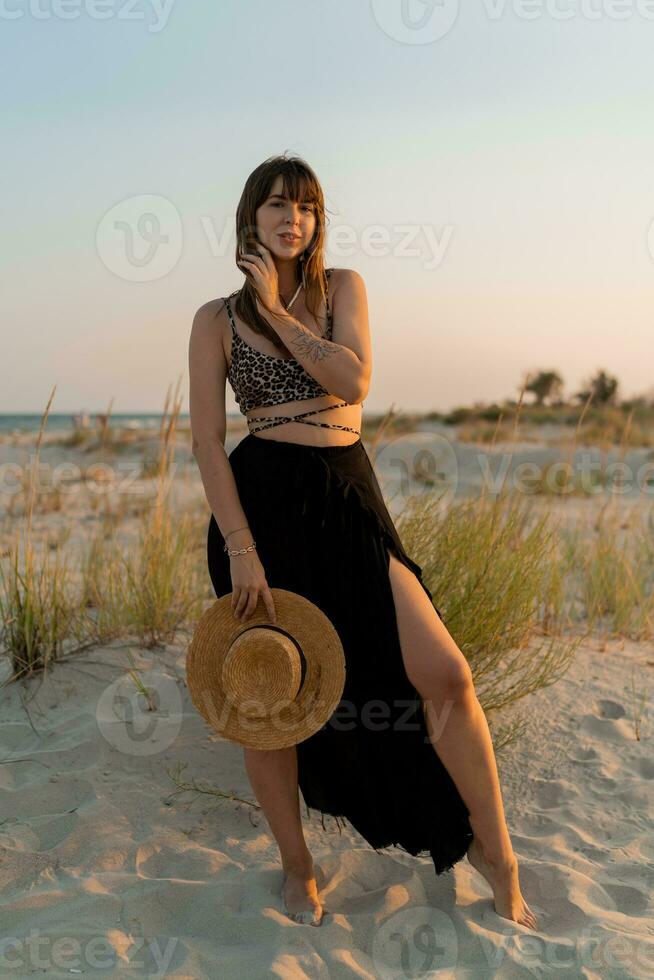 allegro brunetta donna nel elegante estate boho attrezzatura in posa su il spiaggia. tramonto colori. foto