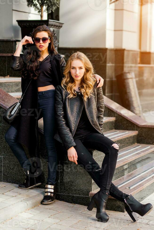 Due elegante bella ragazze in posa su il strada nel soleggiato giorno. indossare di moda urbano attrezzatura , pelle giacca e stivali tacchi. foto