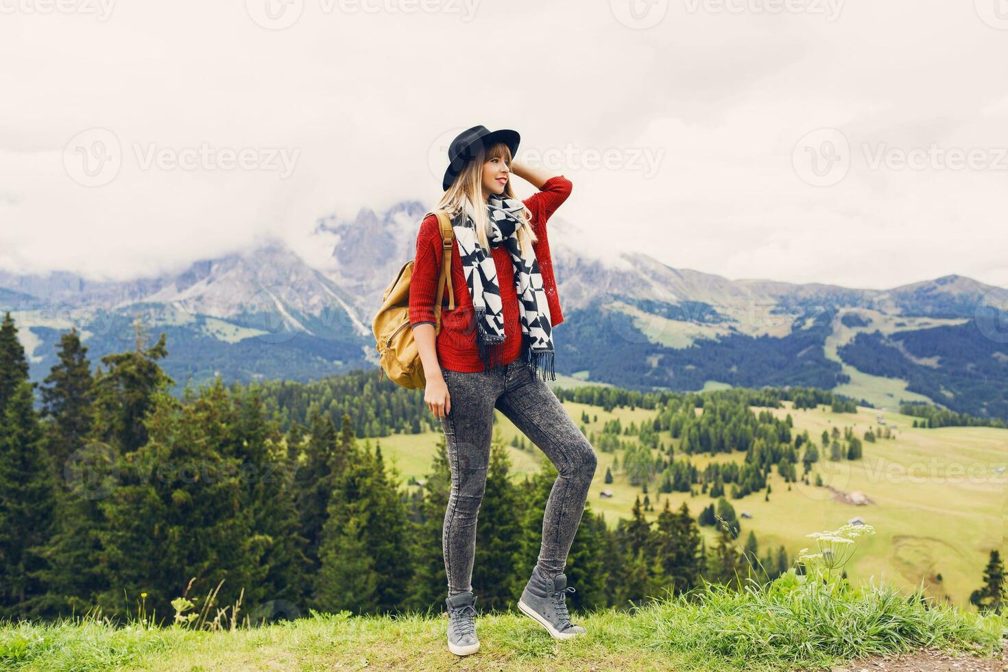 giovane viaggiatore donna con cappello e zaino godendo sorprendente montagna Visualizza ,a passeggio e Esplorare vero natura . ventoso tempo atmosferico. indossare elegante boho autunno vestito, rosso a maglia maglione e jeans. foto