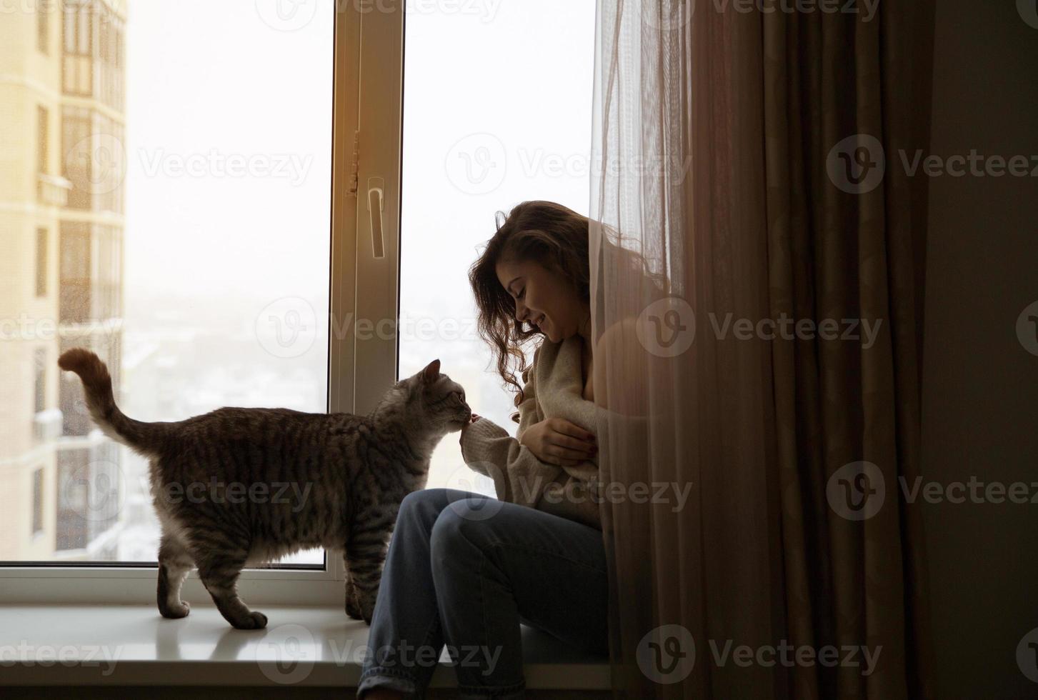 ragazza si siede vicino alla finestra accanto a un gatto foto