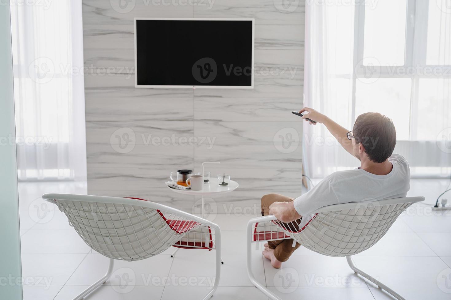 uomo seduto su una sedia che guarda la tv con in mano il telecomando foto