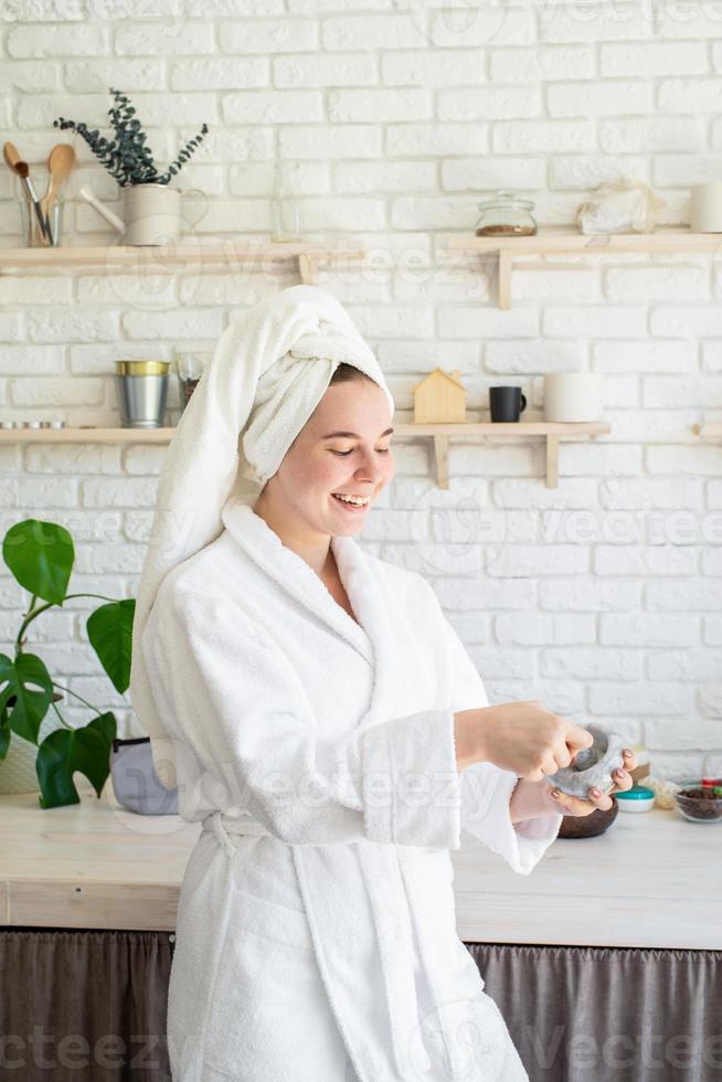 felice giovane donna che applica uno scrub per il viso sul viso nella sua cucina di casa foto
