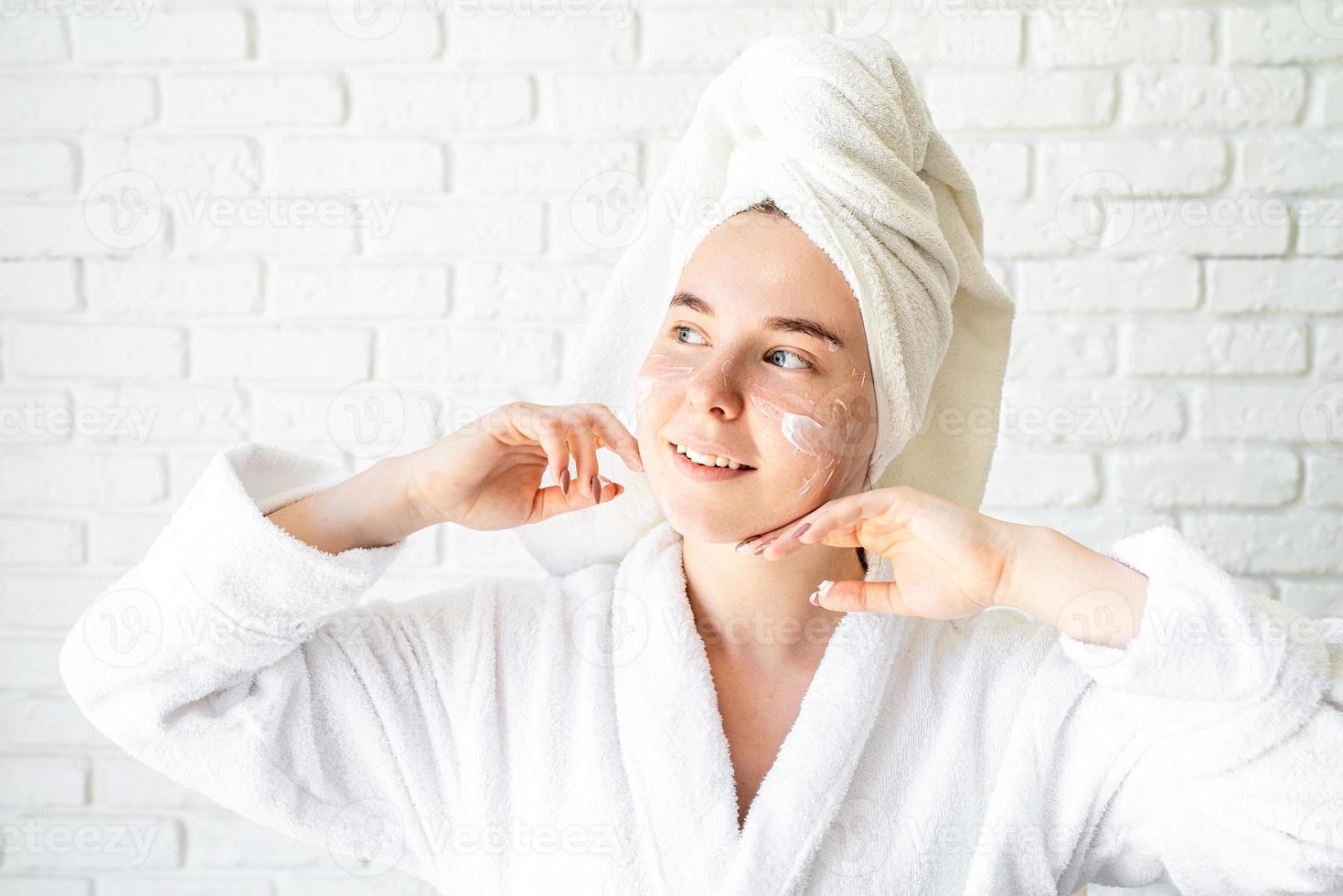 donna caucasica in asciugamano da bagno bianco che applica crema per il viso a casa foto