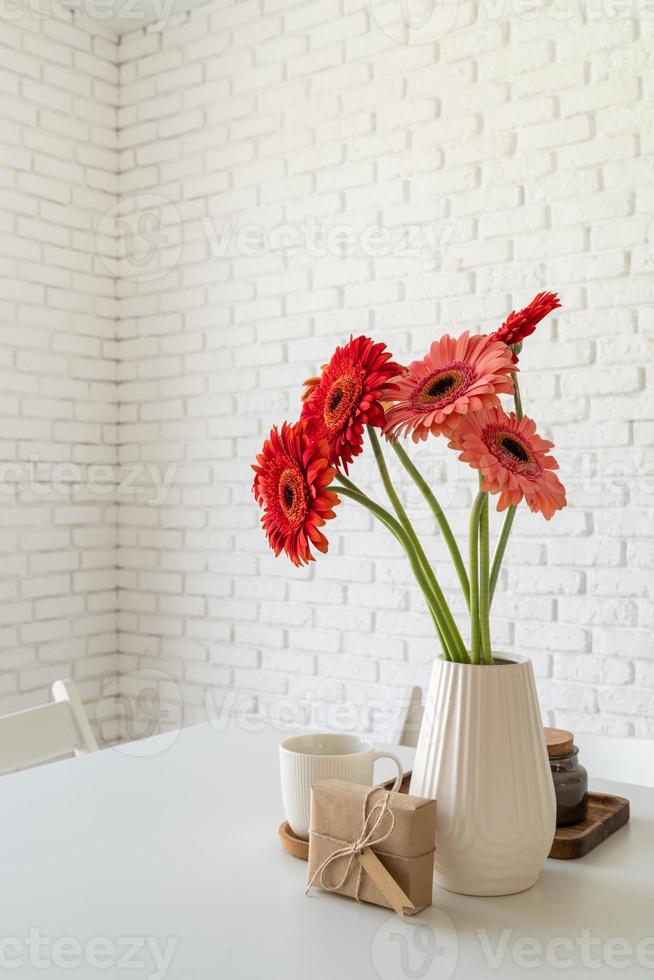 margherite gerbera luminose in vaso bianco sul tavolo della cucina con una confezione regalo foto