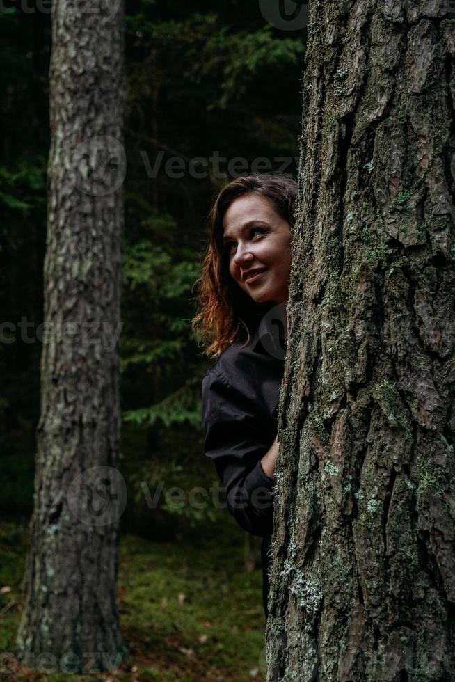 una giovane donna in un bosco di conifere si affaccia da dietro un albero foto