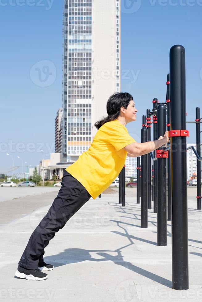 Sorridente donna anziana facendo push up all'aperto sulle barre del campo sportivo foto