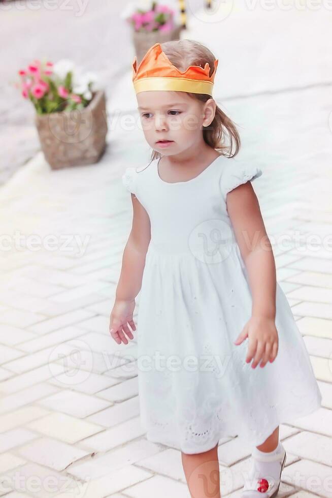 poco bellissimo ragazza nel un' bianca vestito e nel un' corona su sua testa. il bambino passeggiate su il piastrella vicino fiore letti con fiori. foto
