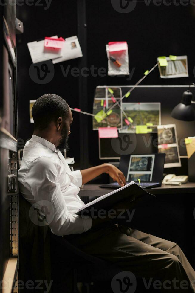 poliziotto stuying crimine Astuccio e analizzando fotografie su computer portatile, seduta a scrivania con prova schema tavola su parete. investigatore controllo sorveglianza telecamera fotografie su computer